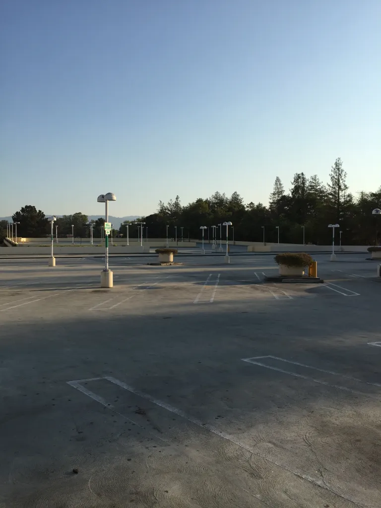 Empty Vallco Mall rooftop parking lot in Cupertino with parking spaces, light poles, trees, and distant hills